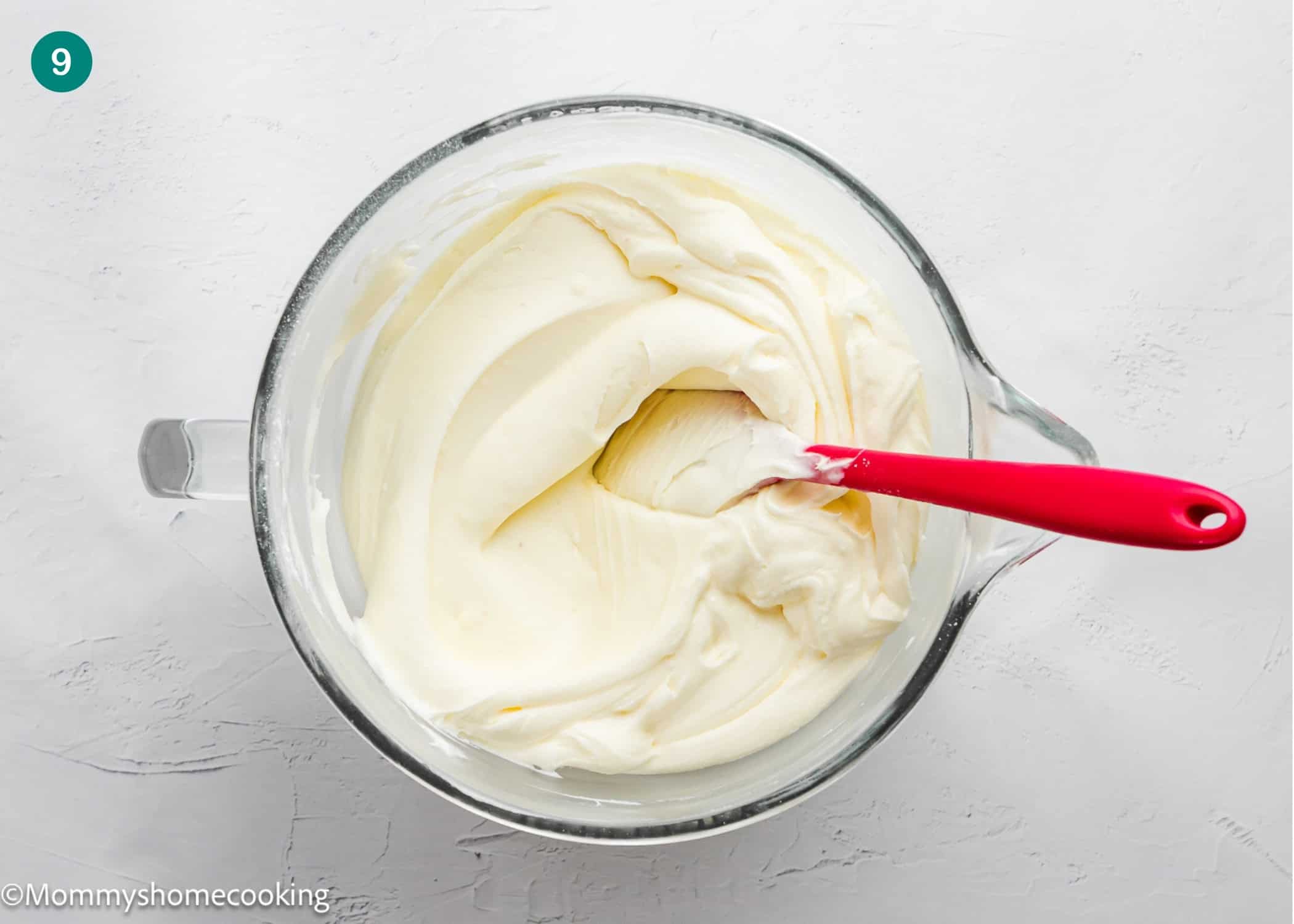 A bowl of creamy white mixture for Eggless Italian Cream Cake with a red spatula resting inside, shown from above on a light textured surface.