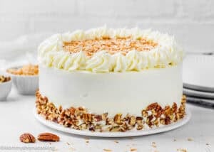 An Eggless Italian Cream Cake with white frosting is topped with toasted coconut flakes and decorated with chopped pecans around the base, sitting elegantly on a white plate.