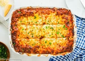 A baked lasagna sliced into twelve pieces in a white dish, topped with melted cheese and sprinkled with chopped parsley, on a table with a blue checkered napkin.