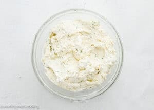 A clear glass bowl filled with a creamy, white cheese mixture with visible green herb flecks, placed on a white textured surface.