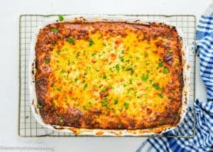 A baked lasagna in a rectangular dish, topped with melted cheese and chopped parsley, sits on a cooling rack next to a blue and white checkered cloth.