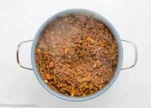 A pot of cooked ground beef mixed with chopped onions, viewed from above on a white surface. Steam rises from the mixture.