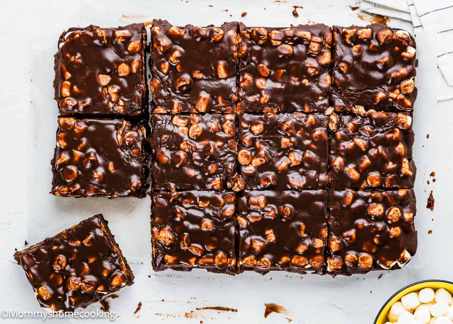 Eggless Brownie squares topped with glossy chocolate and mini marshmallows are arranged on a white surface, with one piece slightly separated from the rest.