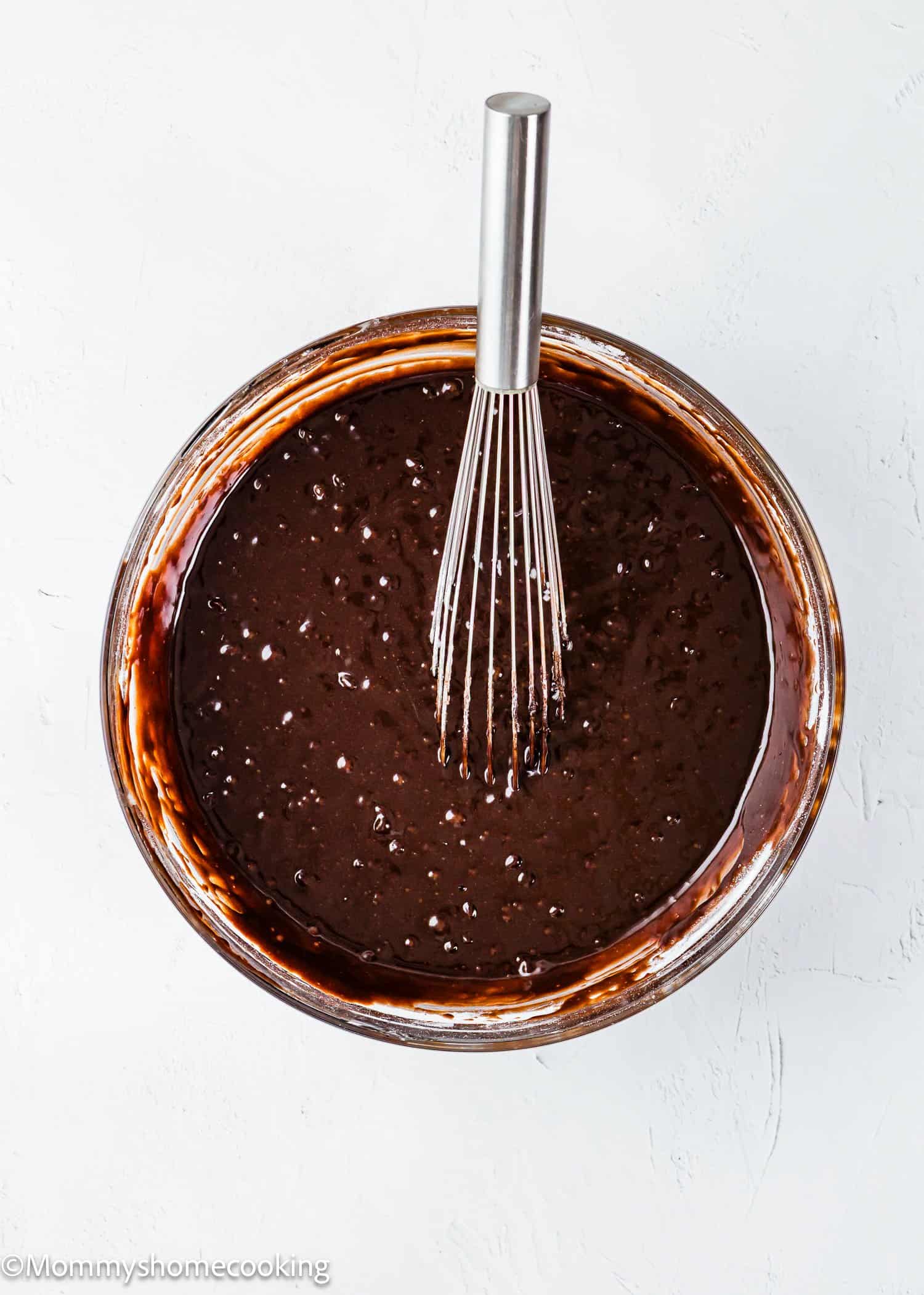 A glass bowl of egg-free chocolate brownie batter with a metal whisk resting inside, set on a white surface.