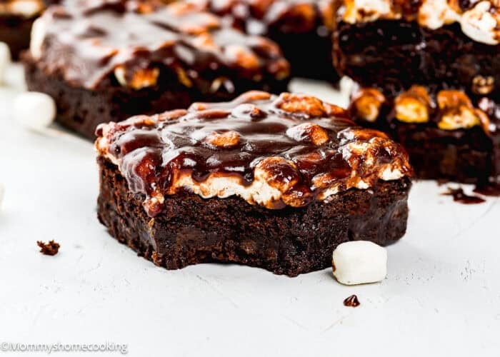 A close-up of a eggless chocolate brownie topped with toasted marshmallows and chocolate sauce, with a bite taken out and a mini marshmallow nearby.