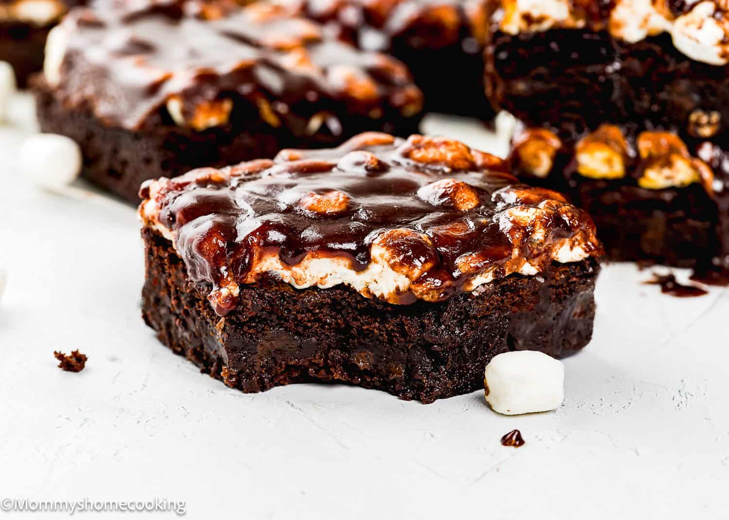 A close-up of a eggless chocolate brownie topped with toasted marshmallows and chocolate sauce, with a bite taken out and a mini marshmallow nearby.