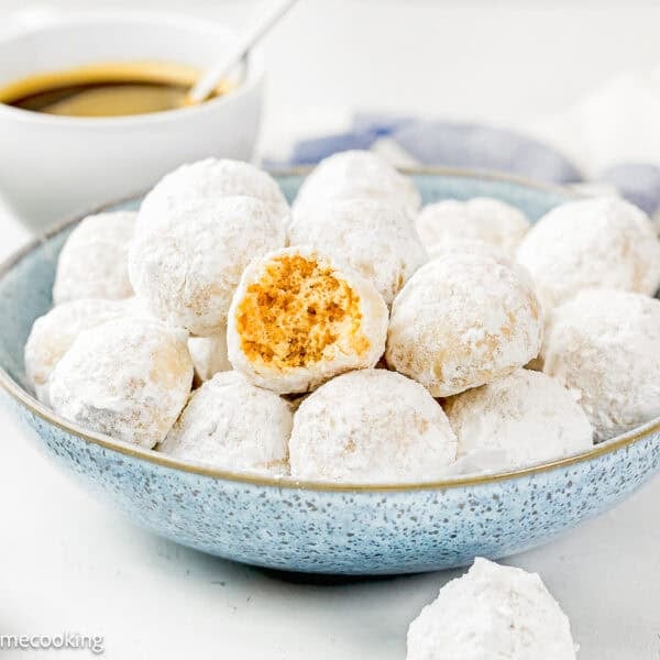 A blue bowl holds Eggless Mini Powdered Sugar Donut Muffins, one with a bite taken out, on a white surface with a cup of coffee in the background.