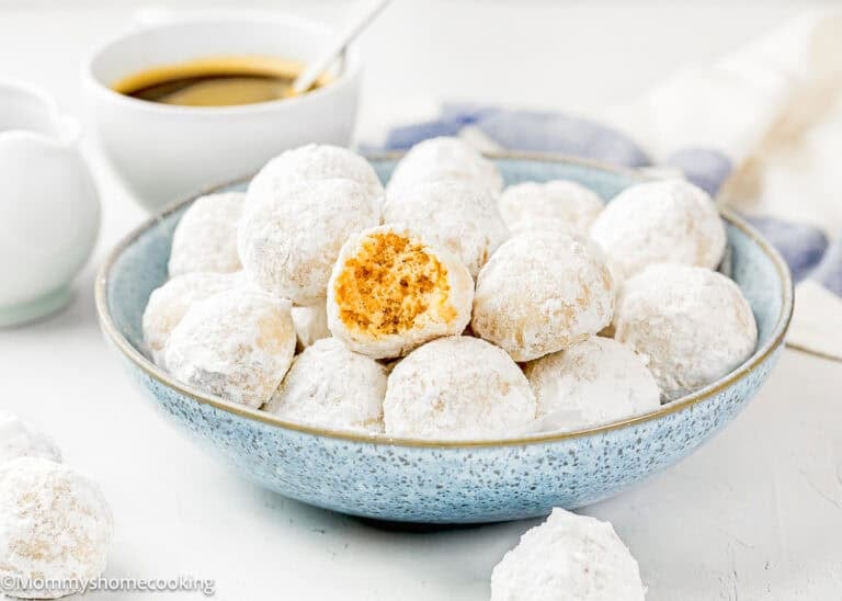 A blue bowl holds Eggless Mini Powdered Sugar Donut Muffins, one with a bite taken out, on a white surface with a cup of coffee in the background.