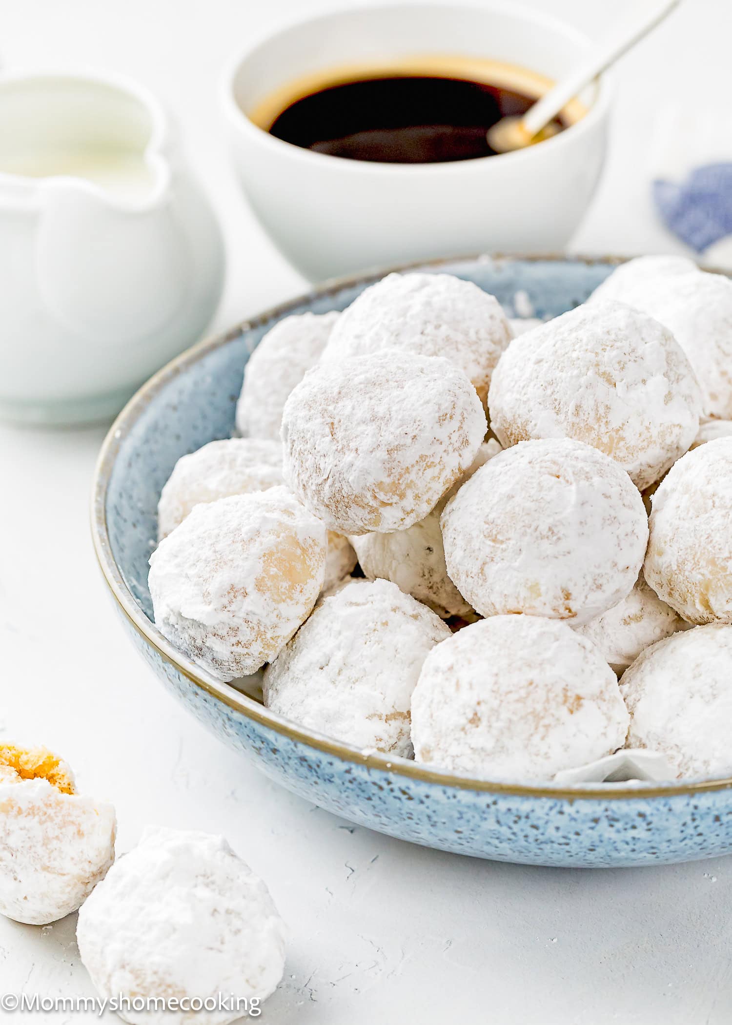 A blue bowl filled with Eggless Mini Powdered Sugar Donut Muffins sits on a white surface, with a cup of black coffee and a small pitcher of milk in the background.