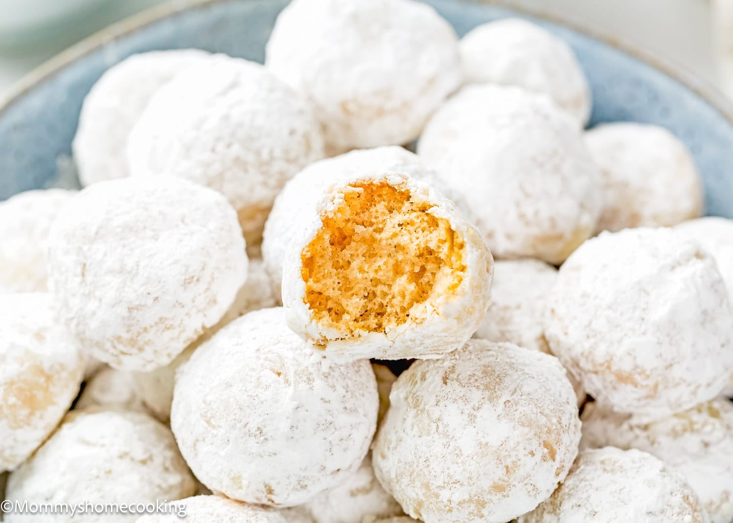 A plate of Eggless Mini Powdered Sugar Donut Muffins, each round and coated in powdered sugar, with one showing a bite taken out to reveal its soft interior.
