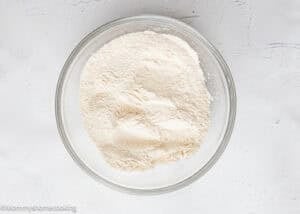 A glass bowl filled with a mound of white flour sits on a textured white surface, ready to make Eggless Mini Powdered Sugar Donut Muffins.