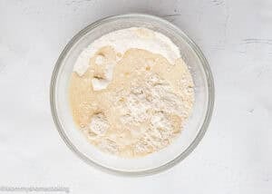 A glass bowl containing a mixture for Eggless Mini Powdered Sugar Donut Muffins, with flour and liquid ingredients partially combined, sits on a white surface.