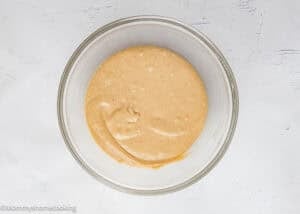 A glass bowl filled with light brown Eggless Mini Powdered Sugar Donut Muffins batter sits on a white textured surface.