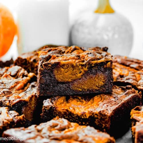 A close-up of several eggless pumpkin brownies stacked on a plate, with a glass of milk and a small pumpkin in the background.
