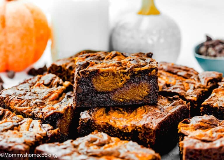 A close-up of several eggless pumpkin brownies stacked on a plate, with a glass of milk and a small pumpkin in the background.