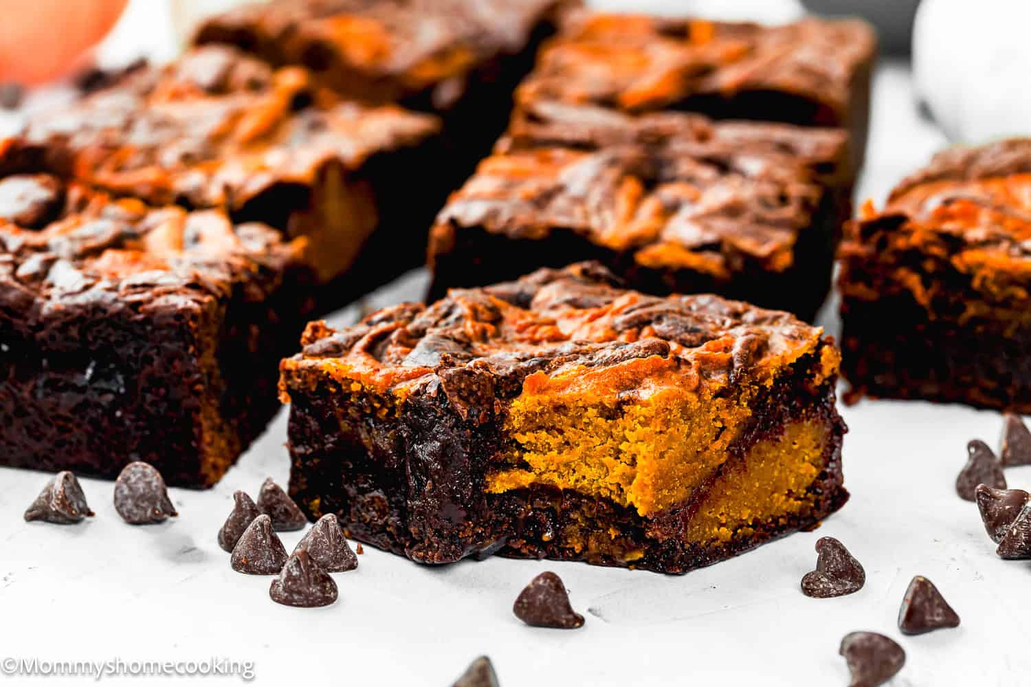 A close-up of eggless pumpkin brownies with a pumpkin swirl, surrounded by scattered chocolate chips on a white surface.