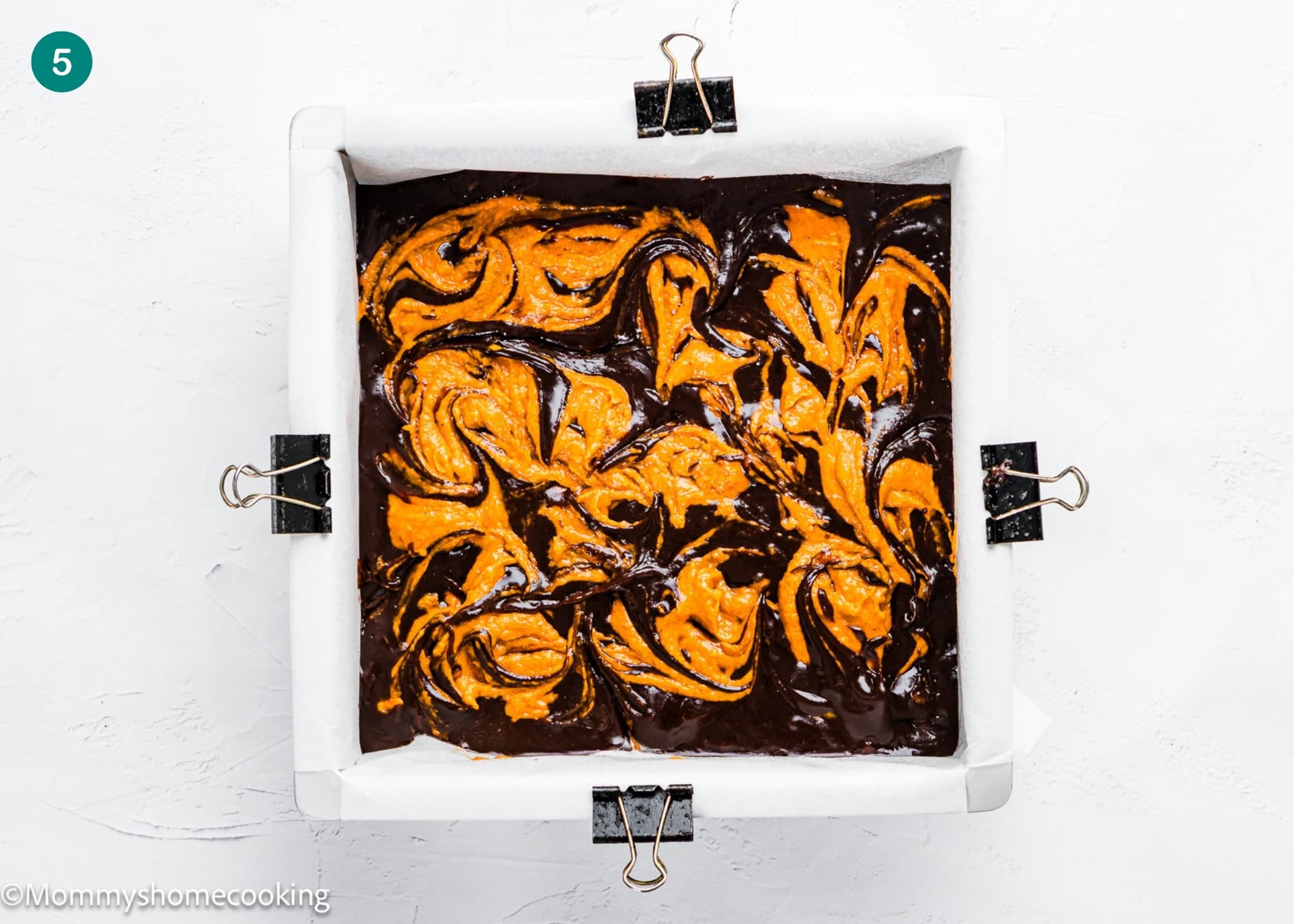 A square baking pan lined with parchment paper holds Eggless Pumpkin Brownies batter, featuring swirls of orange-colored mixture on top and secured with black binder clips.
