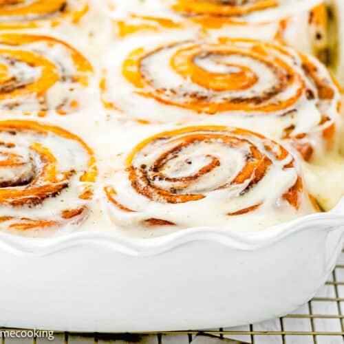 A close-up of eggless pumpkin cinnamon rolls in a white baking dish, topped with a layer of white icing.
