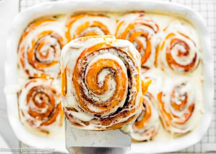 A close-up of an eggless pumpkin cinnamon roll being lifted from a baking dish filled with other glazed cinnamon rolls.