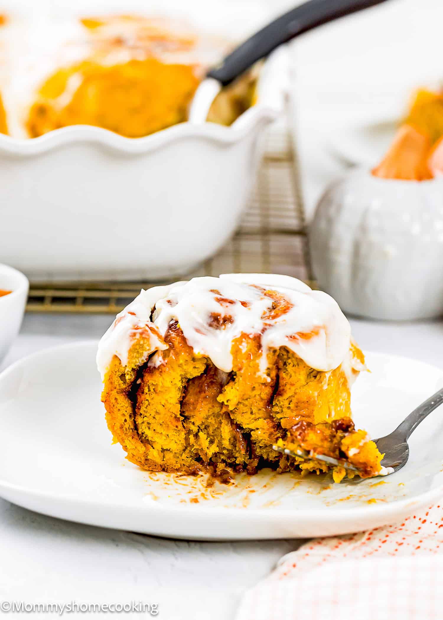 An Eggless Pumpkin Cinnamon Roll topped with cream cheese frosting sits on a white plate with a fork, surrounded by a baking dish and pumpkin decor.