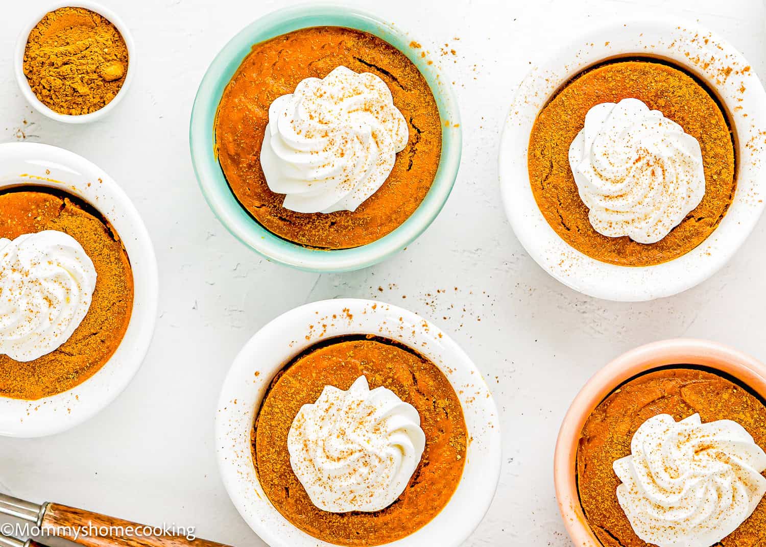 Several small bowls of Eggless Pumpkin Custard Cups (Crustless Pumpkin Pie) topped with whipped cream are arranged on a white surface, with a small bowl of ground cinnamon nearby.