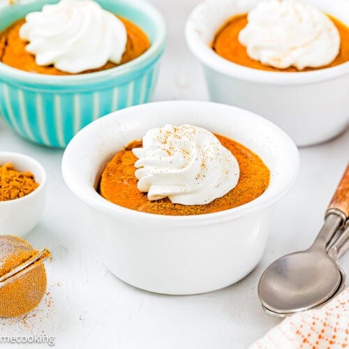Three ramekins of Eggless Pumpkin Custard Cups (Crustless Pumpkin Pie) topped with whipped cream are on a white surface, with a small bowl of ground spice, a mesh strainer, and two spoons nearby.