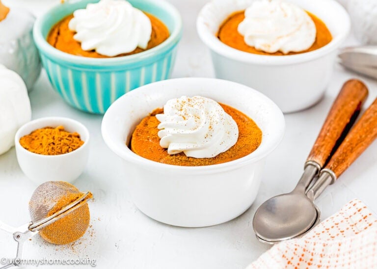 Three ramekins of Eggless Pumpkin Custard Cups (Crustless Pumpkin Pie) topped with whipped cream are on a white surface, with a small bowl of ground spice, a mesh strainer, and two spoons nearby.