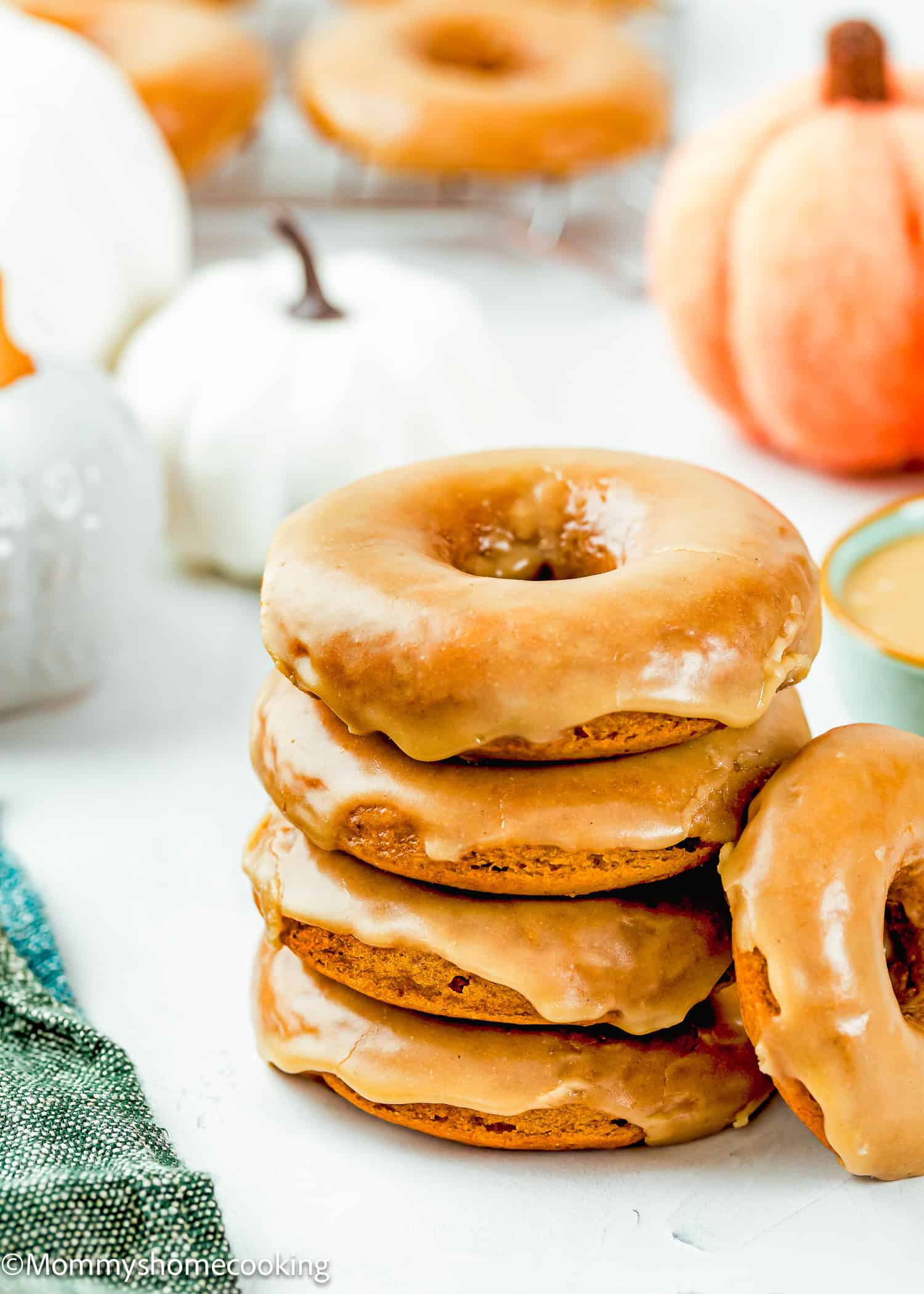 A stack of four Eggless Pumpkin Donuts sits on a white surface with pumpkins and more donuts in the background.