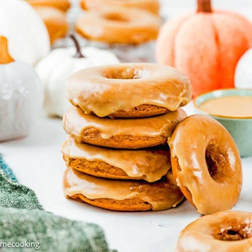 A stack of Eggless Pumpkin Donuts is displayed on a white surface with more donuts, ceramic pumpkins, a green napkin, and a bowl of glaze in the background.