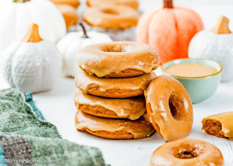 A stack of Eggless Pumpkin Donuts is displayed on a white surface with more donuts, ceramic pumpkins, a green napkin, and a bowl of glaze in the background.