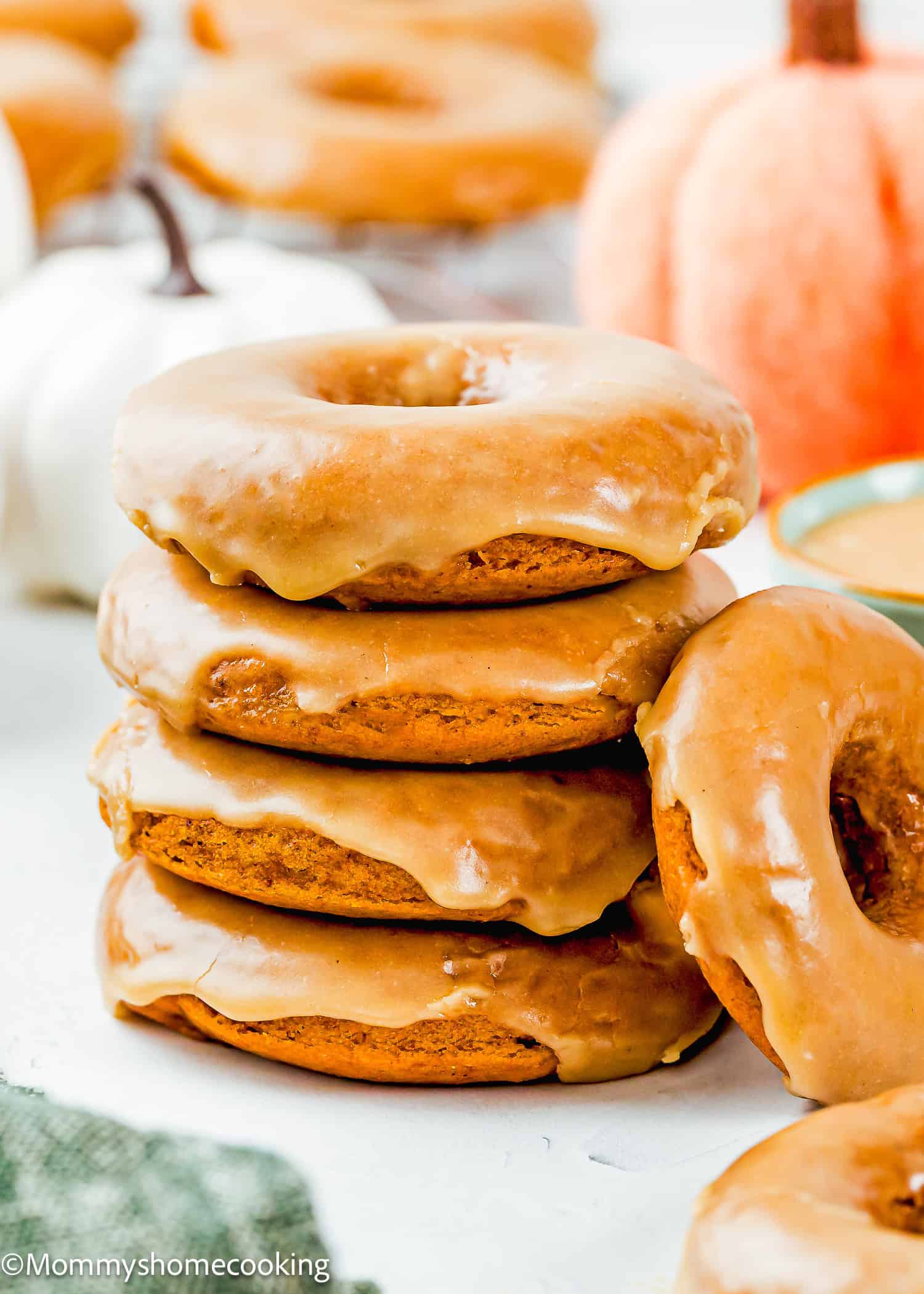 A stack of four eggless pumpkin donuts with light brown frosting sits on a white surface, with a pumpkin and more doughnuts in the background.