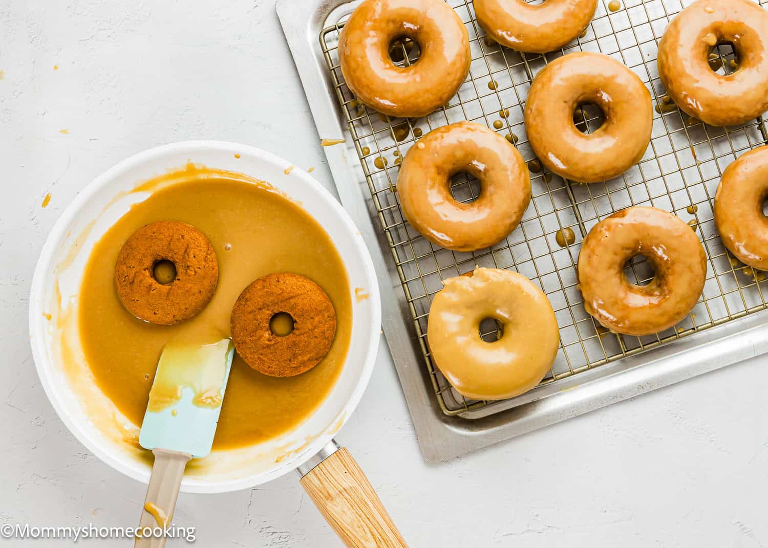 A baking tray with Eggless Pumpkin Donuts on a wire rack, next to a bowl of caramel glaze where two donuts are being dipped with a spatula.