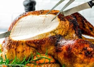 A golden roasted turkey is being carved with a knife and fork, revealing tender white meat inside. This is not a classic boring turkey: Citrus-Garlic Chipotle Thanksgiving Turkey Recipe, served with fresh rosemary and vegetables underneath.