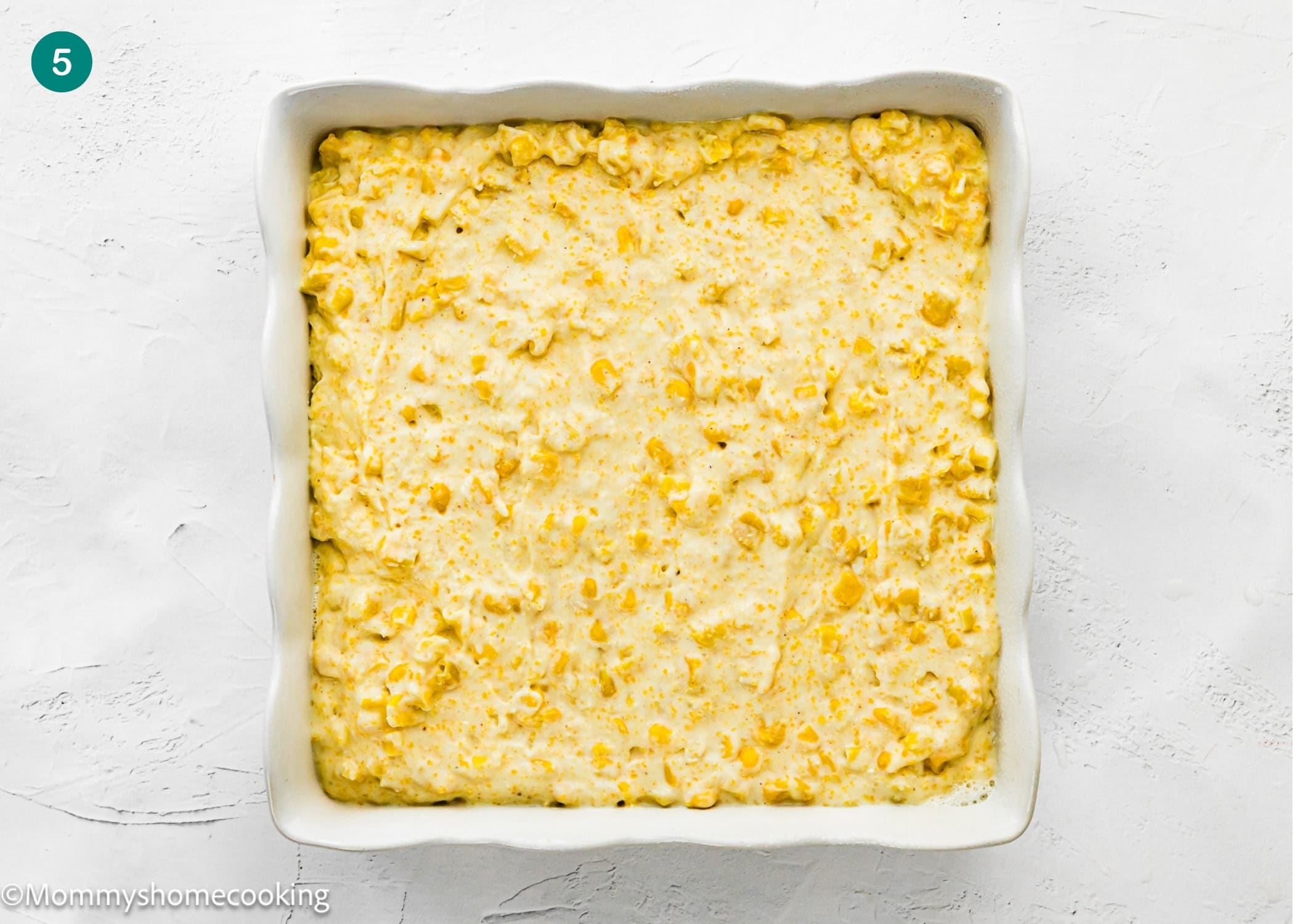 A square white baking dish filled with an unbaked Easy Homemade Corn Pudding without Eggs sits on a white surface.