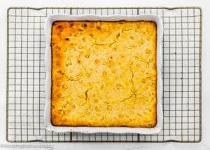 A square baking dish filled with golden-brown Easy Homemade Corn Pudding without Eggs sits on a cooling rack.
