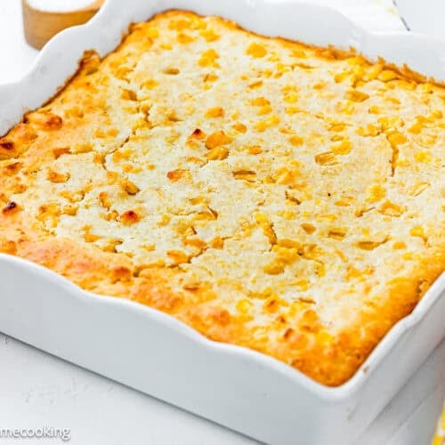 An easy homemade corn pudding without eggs sits golden-brown in a white rectangular dish on a white surface, with a checkered cloth partially visible beside it.