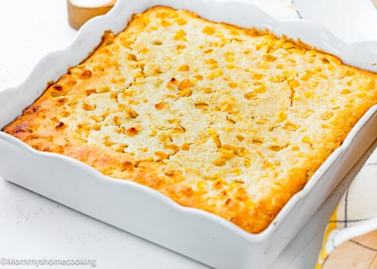An easy homemade corn pudding without eggs sits golden-brown in a white rectangular dish on a white surface, with a checkered cloth partially visible beside it.