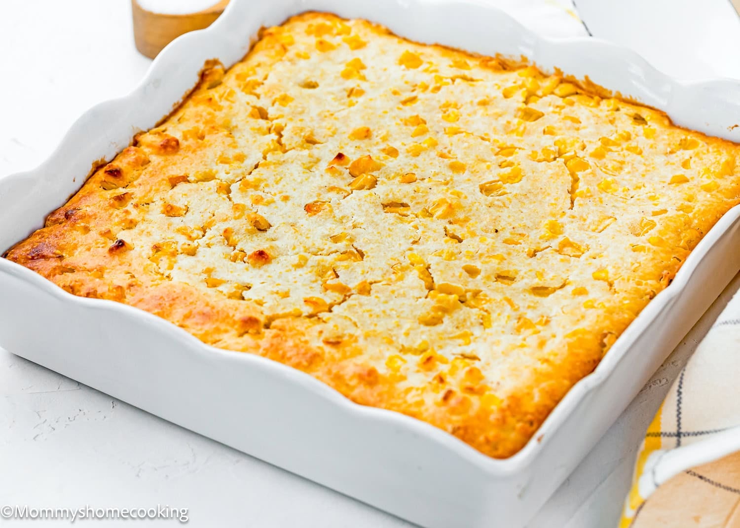 An easy homemade corn pudding without eggs sits golden-brown in a white rectangular dish on a white surface, with a checkered cloth partially visible beside it.