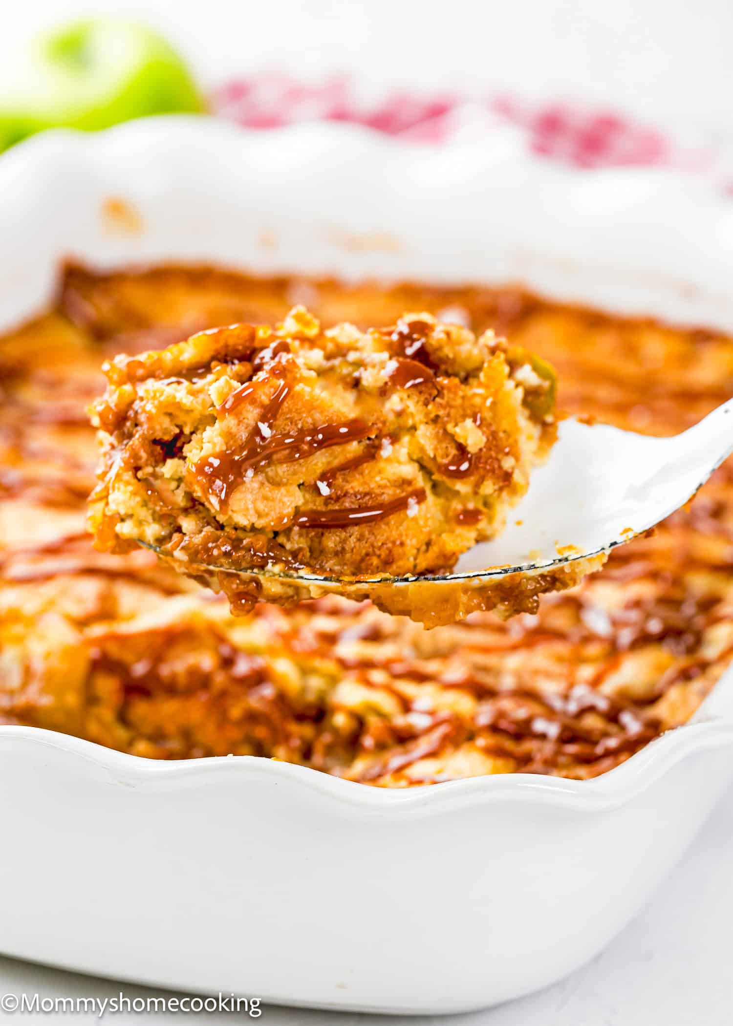 A close-up of a serving spoon lifting a portion of Eggless Apple Caramel Dump Cake from a white baking dish.
