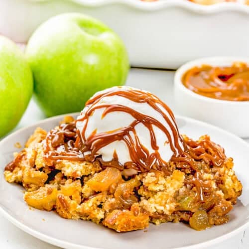 A plate of Eggless Apple Caramel Dump Cake topped with vanilla ice cream and caramel drizzle, with green apples and caramel sauce in the background.