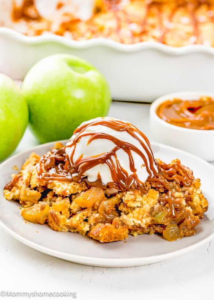 A plate of Eggless Apple Caramel Dump Cake topped with vanilla ice cream and caramel drizzle, with green apples and caramel sauce in the background.