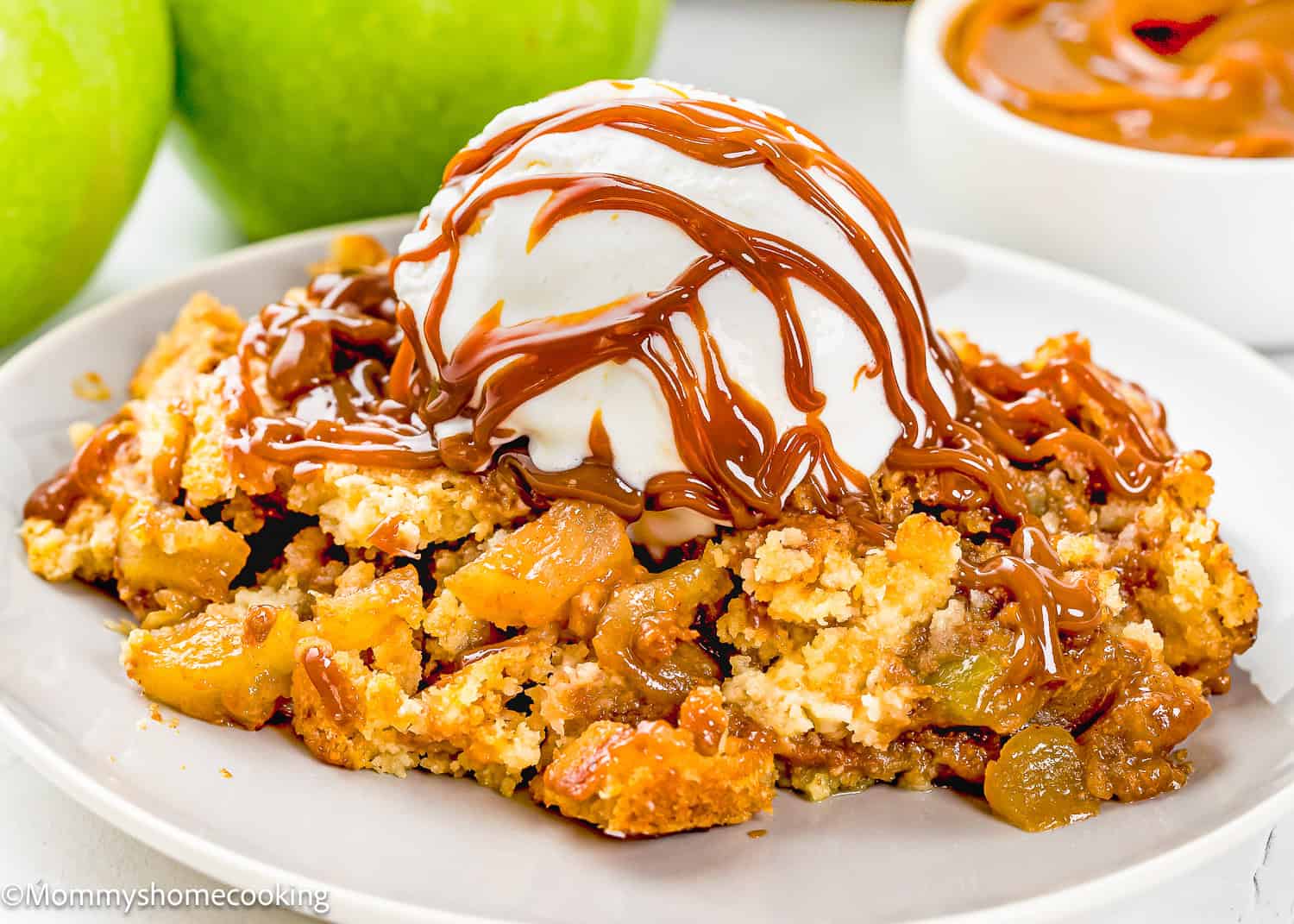 A serving of Eggless Apple Caramel Dump Cake topped with a scoop of vanilla ice cream and drizzled with caramel sauce on a white plate.