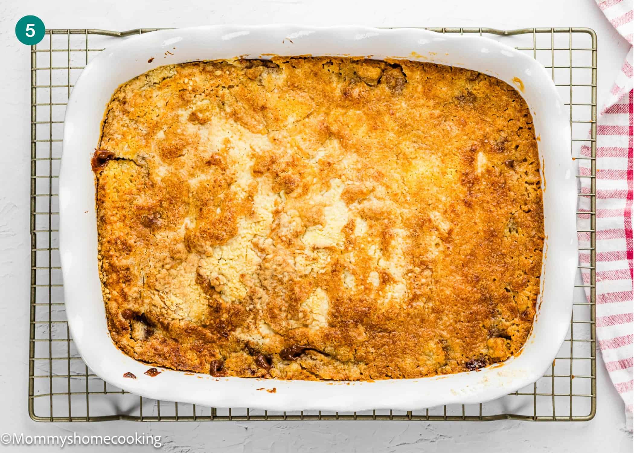 An Eggless Apple Caramel Dump Cake with a golden-brown top sits in a white rectangular dish on a cooling rack, with a red-striped towel beside it.