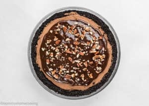 An Eggless Chocolate Mousse Pie (no-bake) with a cookie crust, topped with glossy chocolate ganache and chopped pecans, is beautifully displayed on a white surface.