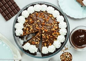 An Eggless Chocolate Mousse Pie (no-bake) with a whipped cream border and chopped nuts on top sits on a table, with a slice cut out and served on a plate nearby.