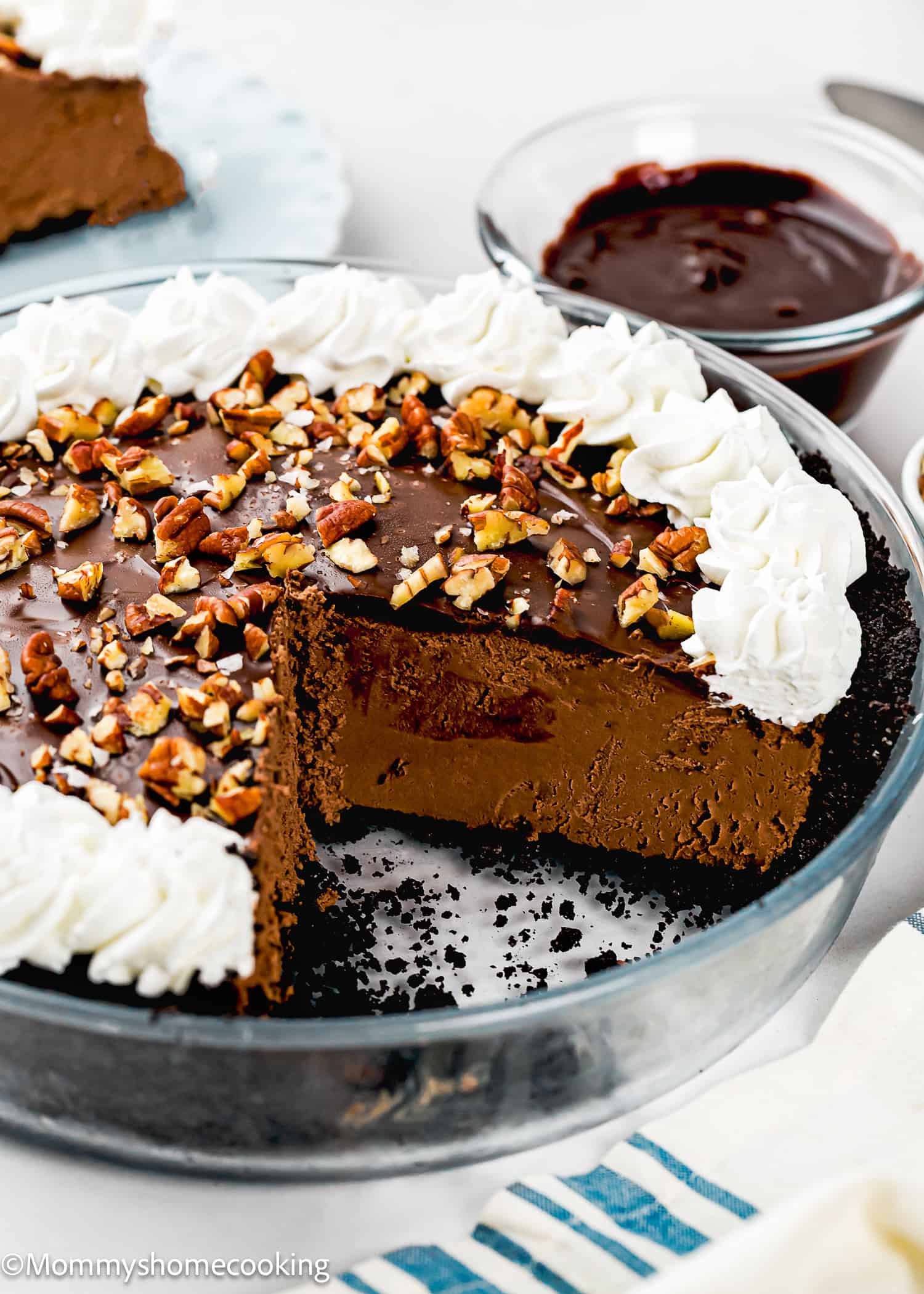 An Eggless Chocolate Mousse Pie (no-bake) with a slice missing is topped with chopped nuts and whipped cream, sitting in a glass pie dish with a bowl of chocolate sauce in the background.