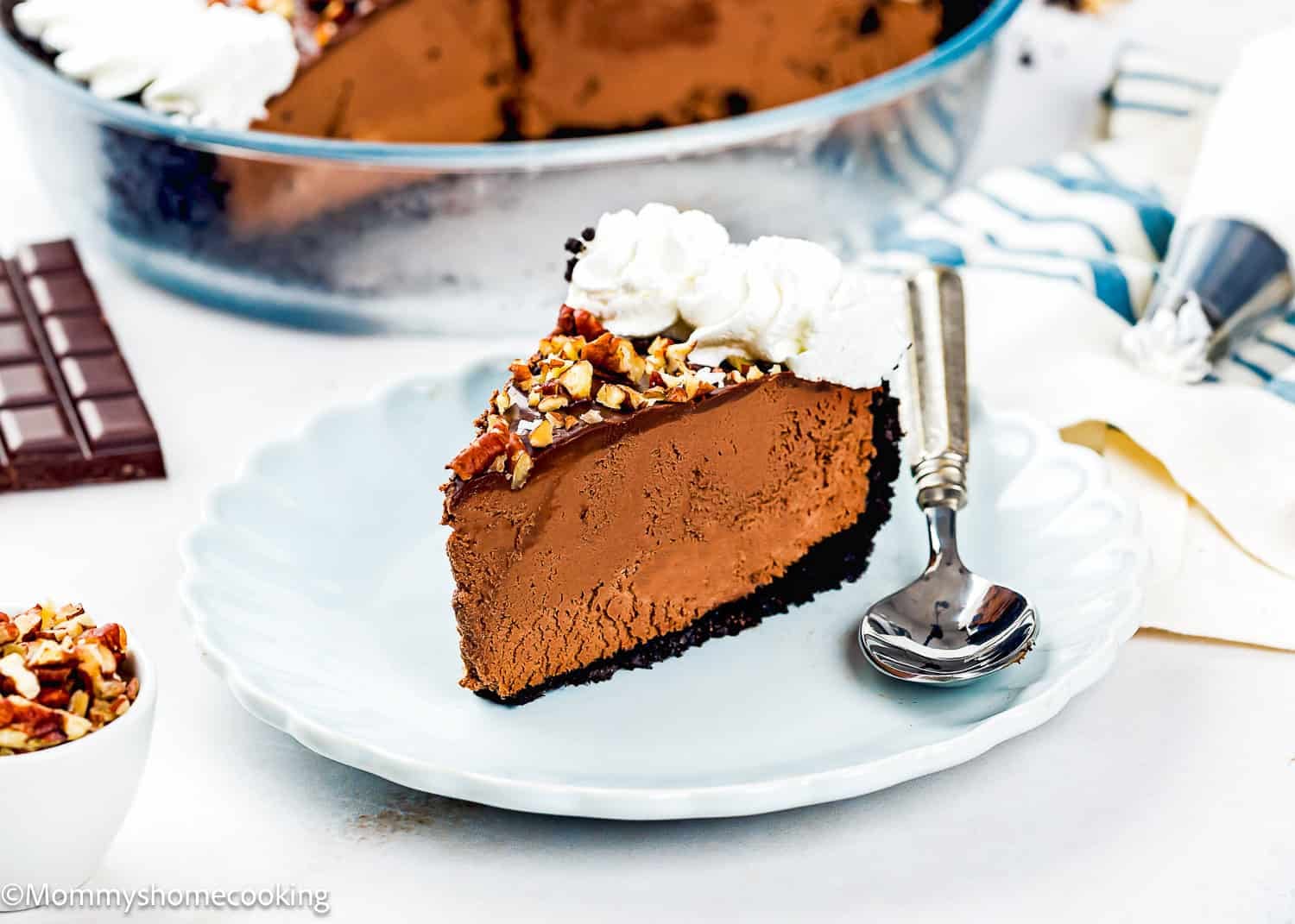 A slice of Eggless Chocolate Mousse Pie (no-bake) with whipped cream and chopped nuts sits on a white plate, a spoon beside it and a pie dish in the background.