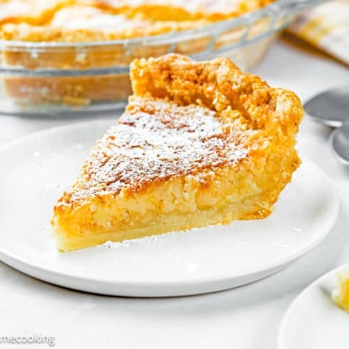 A slice of Eggless Classic Chess Pie Recipe topped with powdered sugar sits on a white plate, with a pie dish and utensils in the background.