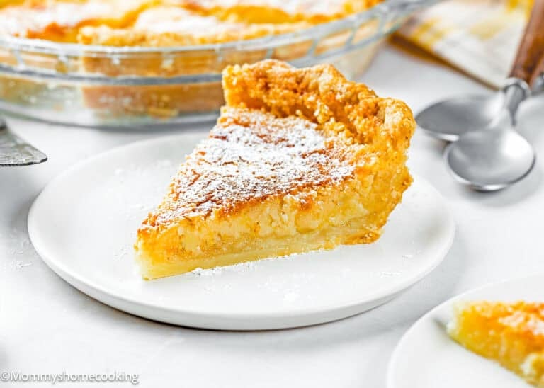 A slice of Eggless Classic Chess Pie Recipe topped with powdered sugar sits on a white plate, with a pie dish and utensils in the background.
