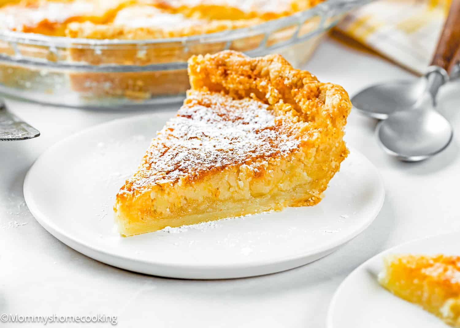 A slice of Eggless Classic Chess Pie Recipe topped with powdered sugar sits on a white plate, with a pie dish and utensils in the background.